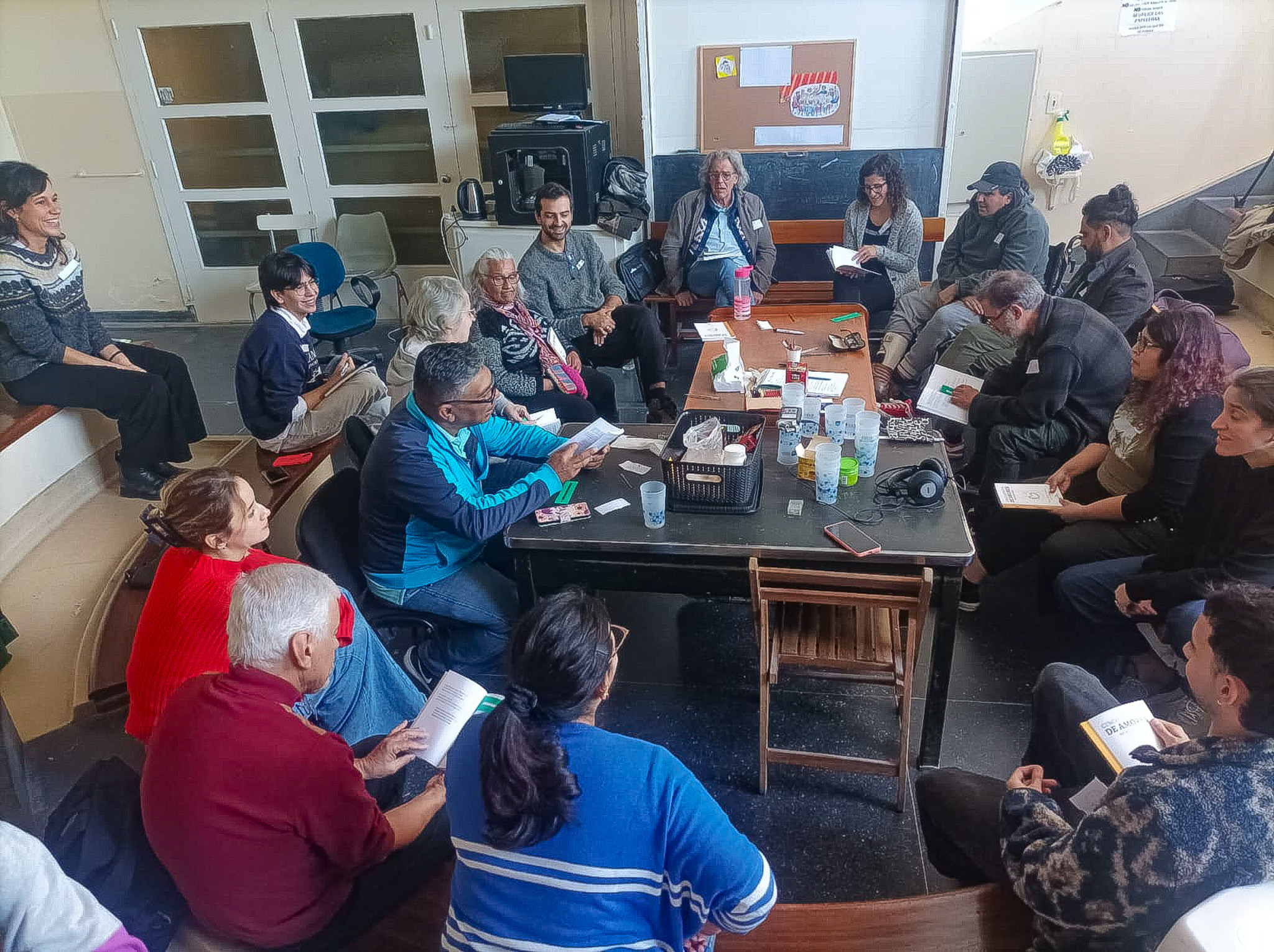 Varias personas con textos sentadas alrededor de mesas con vasos, celulares, entre otros.