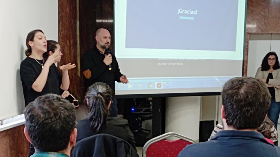 Federico Lezama habla de pie junto a una pantalla en la que se proyecta una presentación. A su lado, una intérprete de LSU. Del lado derecho de la imagen, Leticia Lorier se encuentra de pie, también junto a la presentación que está siendo proyectada.