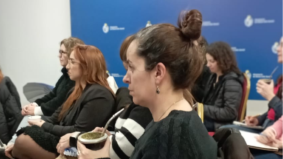 Varias personas sentadas, mirando hacia la izquierda de la imagen. En el centro, una mujer, mira atenta hacia adelante con un mate en la mano.