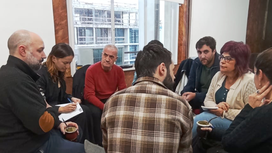 Varias personas sentadas en ronda, conversan. Al centro de la imagen, un hombre de espaldas mira atentamente a su compañera, quien se encuentra a la derecha.