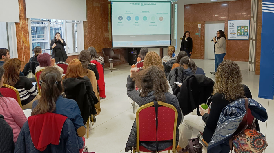 Varias personas sentadas de espalda a la cámara, mirando al frente hacia una presentación proyecada. Del lado derecho de la imagen, Leticia Lorier de pie hablando al micrófono.