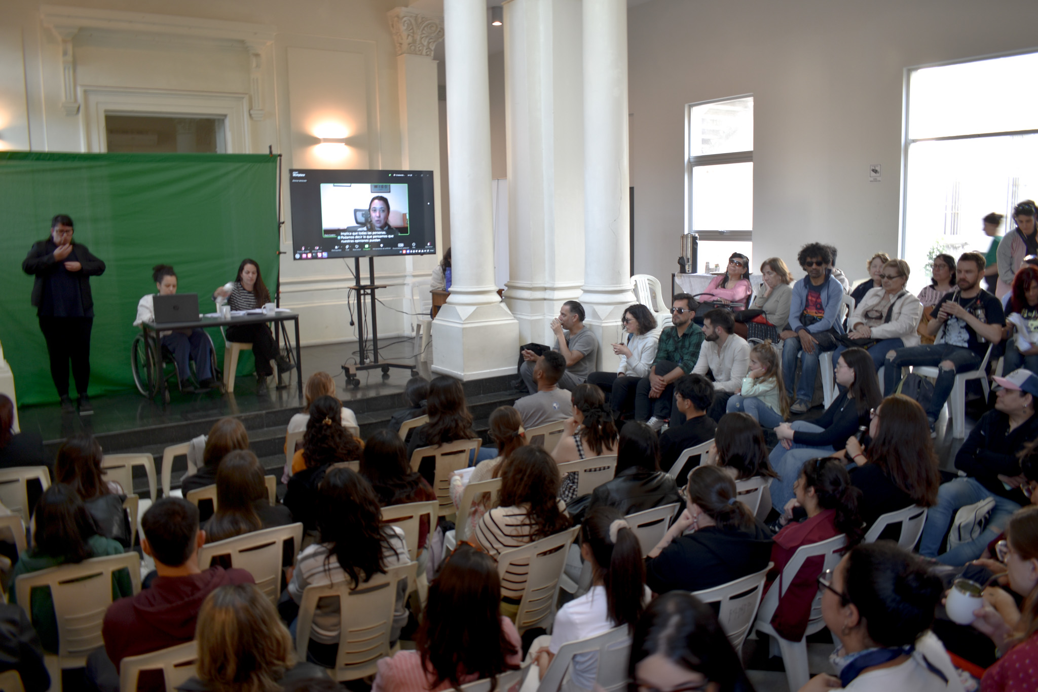 Varias personas en el portal Rabú sentadas en sillas miran hacia adelante. Al frente, una televisión con llamada zoom, con una persona hablando. A la izquierda de la pantalla,