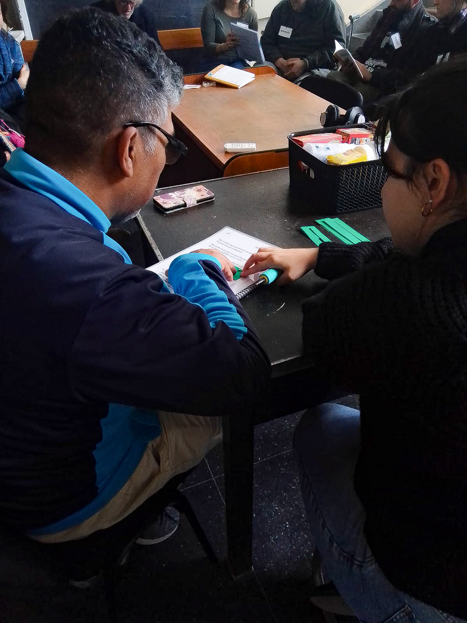 Un hombre y una mujer sentados de espalda a la cámara comparten un texto con una regleta verde sobre el mismo, Sobre la mesa, más regletas y una caja con cosas.