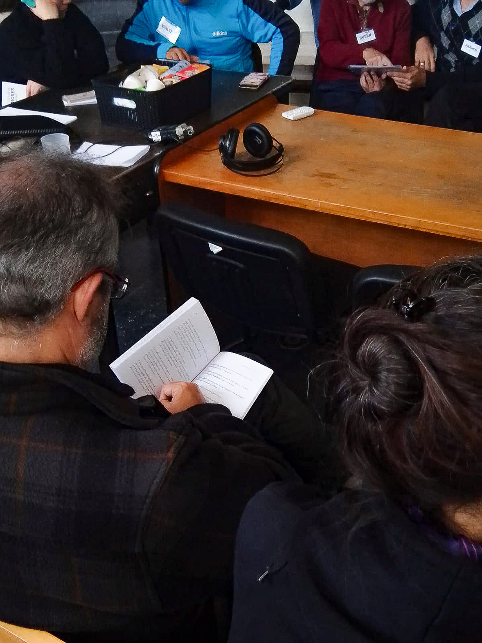Un hombre de espalda con un libro en la mano. Sobre la mesa, unos auriculares y una caja con cosas. Varias personas sentadas alrededor de la mesa.