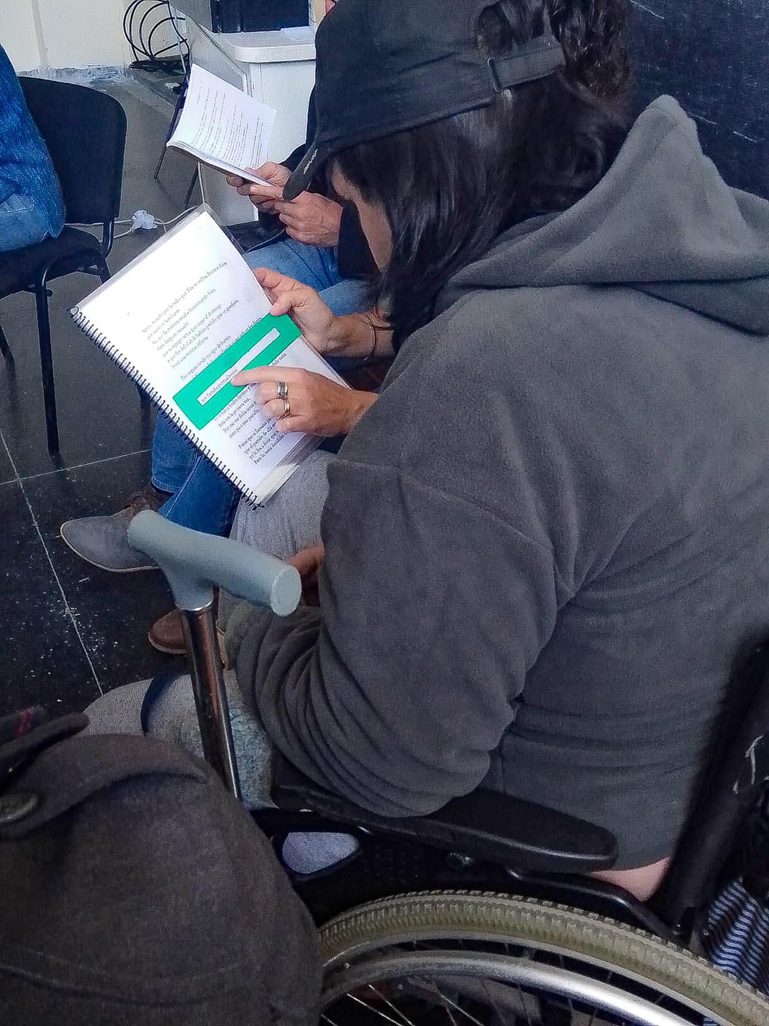 Una persona en silla de ruedas, de espalda a la cámara, está atenta a un cuaderno con una regleta verde colocada. El cuaderno es sostenido por las manos de la persona que está a su lado, quien no sale en la foto.