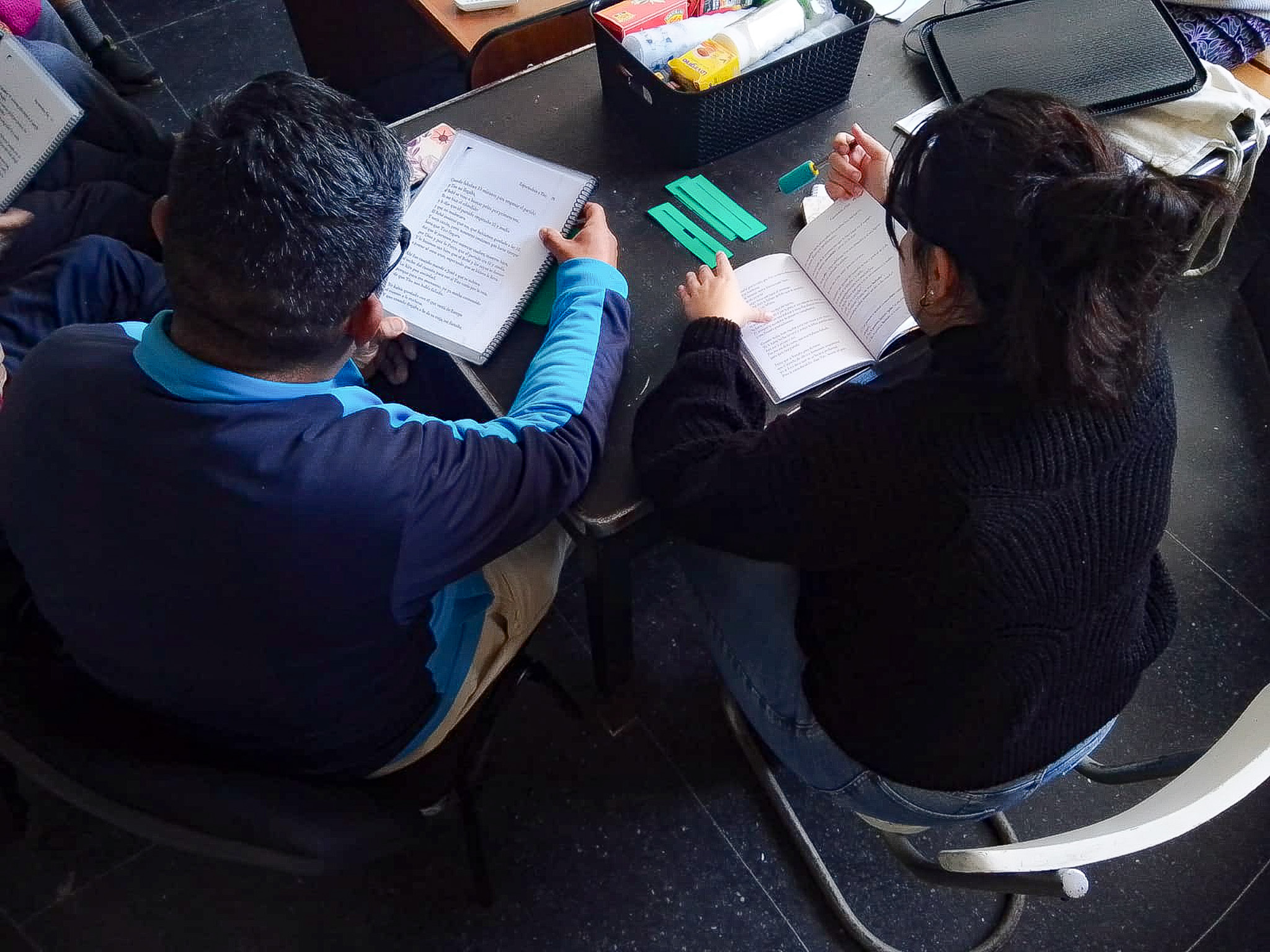 Un hombre y una mujer sentados de espalda a la cámara con textos en la mano. Sobre la mesa, unas regletas de color verde.
