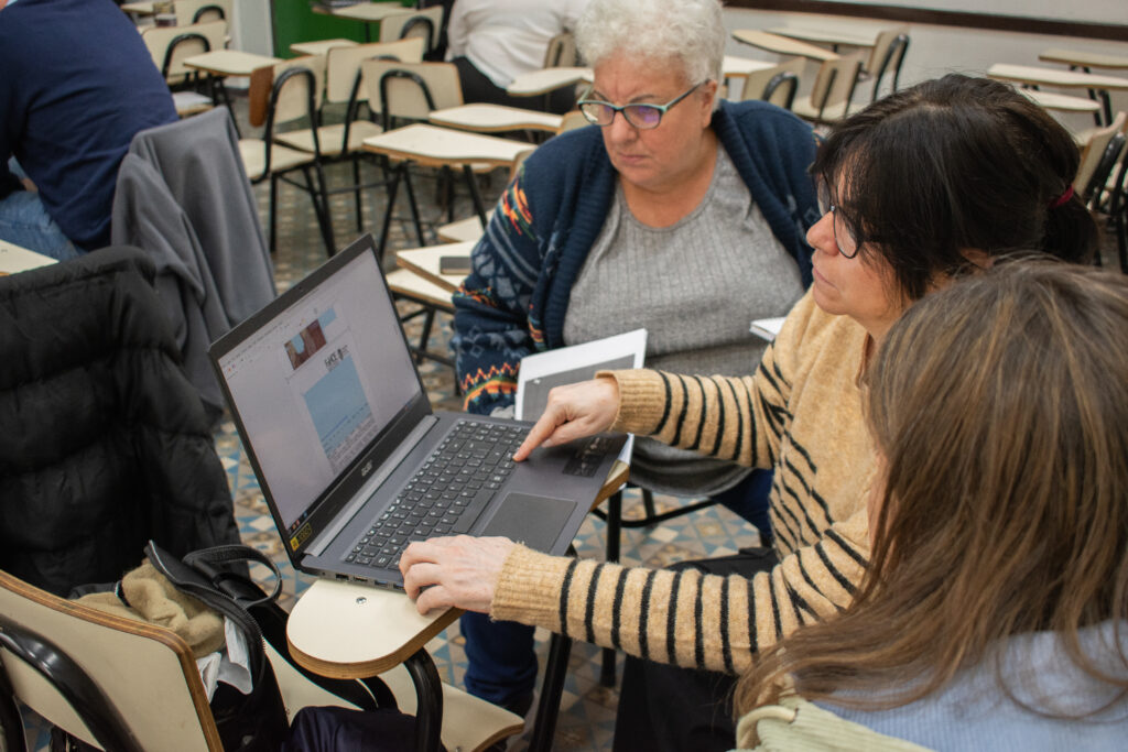 Tres mujeres sentadas en bancos de un salón de clase. Una de ellas, sentada en medio, trabaja con una computadora portatil. Las otras dos mujeres miran atentamente la pantalla de la computadora. 