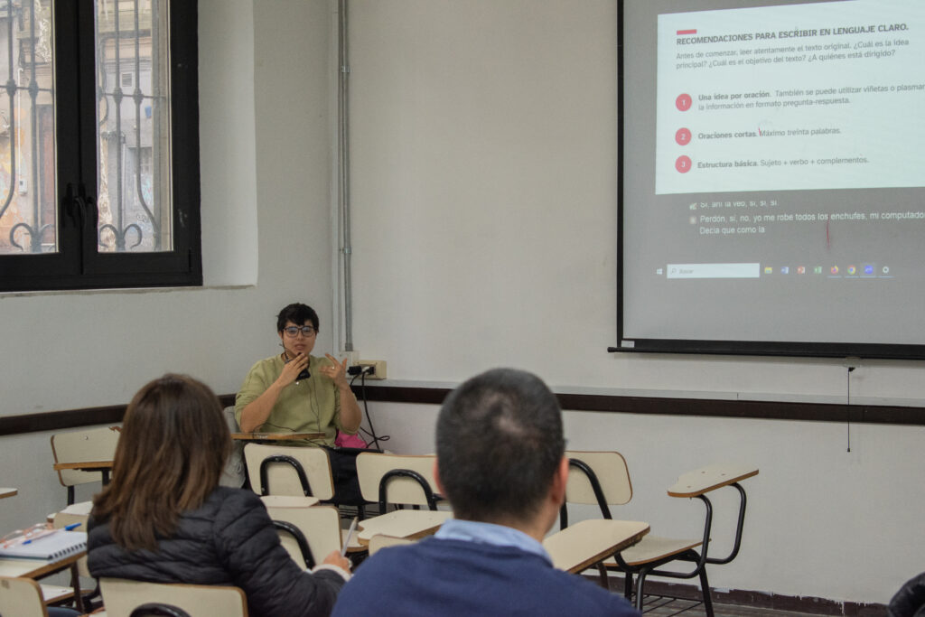 Desde la perspectiva de la clase, dos personas miran hacia la proyección en la pantalla. Belén González mira en dirección a la clase y habla a un dispositivo con cables.