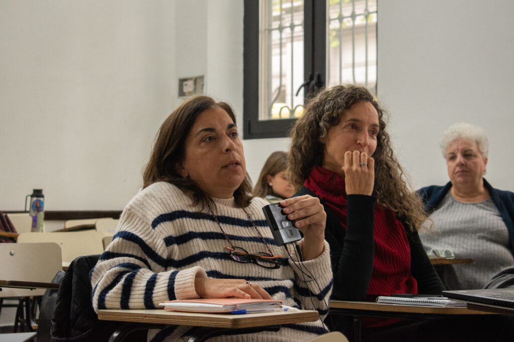Dos mujeres mirando hacia adelante sentadas en bancos de salón. Una de ellas está hablando y sujeta un dispositivo con cables.