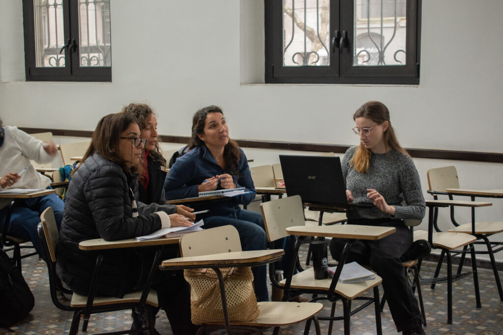 Cuatro mujeres sentadas en  bancos de salón, Una de ellas escribe en una computadora portatil.