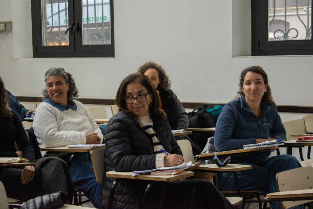 Varias mujeres sentadas en bancos de clase, miran hacia su derecha sonrientes. 