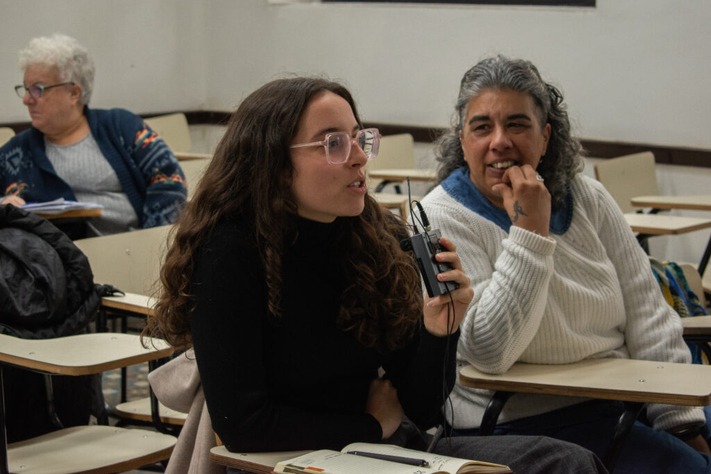 Dos mujeres sentadas en bancos de clase. Una de ellas habla a un dispositivo que sostiene en su mano. La otra mujer la mira sonriente. 