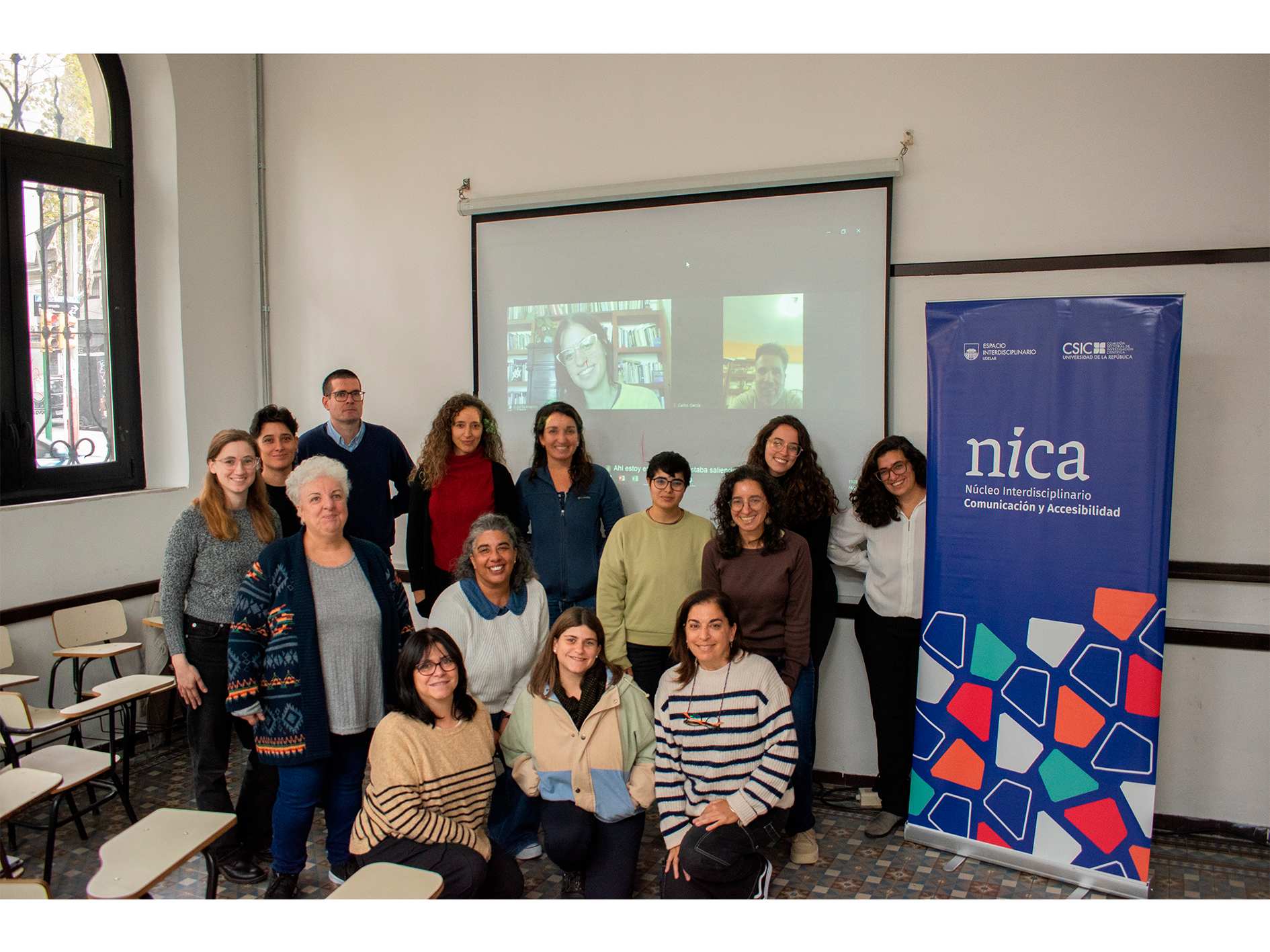 Varias personas posan sonrientes a la cámara. De fondo, una proyección con dos personas particpando via zoom. A la derecha, un banner plegable del NICA